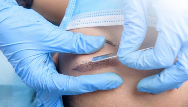 Technician performing a microblading eyebrow treatment on a client wearing a face mask